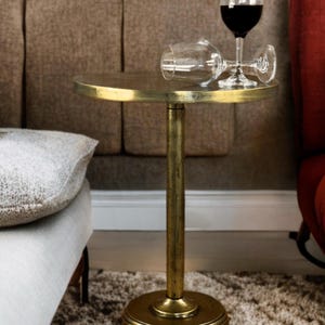 Könnte beinhalten: Ein kleiner, runder Beistelltisch mit goldener Oberfläche. Zwei Weingläser, eines aufrecht und mit Rotwein gefüllt, das andere auf der Seite liegend, stehen auf dem Tisch. Der Tisch hat einen einzelnen Mittelständer und einen kreisförmigen Sockel. Der Hintergrund umfasst ein braunes Sofa und einen gemusterten Teppich.