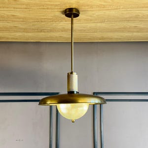 Może przedstawiać: Złota lampa wisząca z kloszem z matowego szkła wisząca z sufitu. Lampa ma cylindryczną podstawę i szeroki, płaski, okrągły klosz.