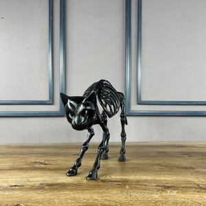 May include: A black cat skeleton ornament, featuring a detailed skeletal design. The cat is depicted in a stalking posture, with its head down and legs slightly flexed. The ornament is made of a glossy black material and rests on a wooden surface.