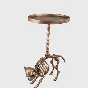 May include: A bronze-colored side table with a cat skeleton design. The table features a round top supported by a bamboo-style column, with the cat skeleton forming the base. A unique decorative accent.