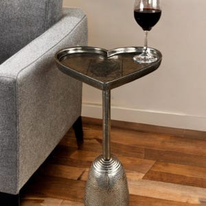 Puede incluir: Una mesa auxiliar en forma de corazón con acabado plateado. La mesa tiene un solo tallo y una base texturizada. Una copa de vino tinto está sobre la superficie de la mesa. La mesa está junto a un sofá gris.