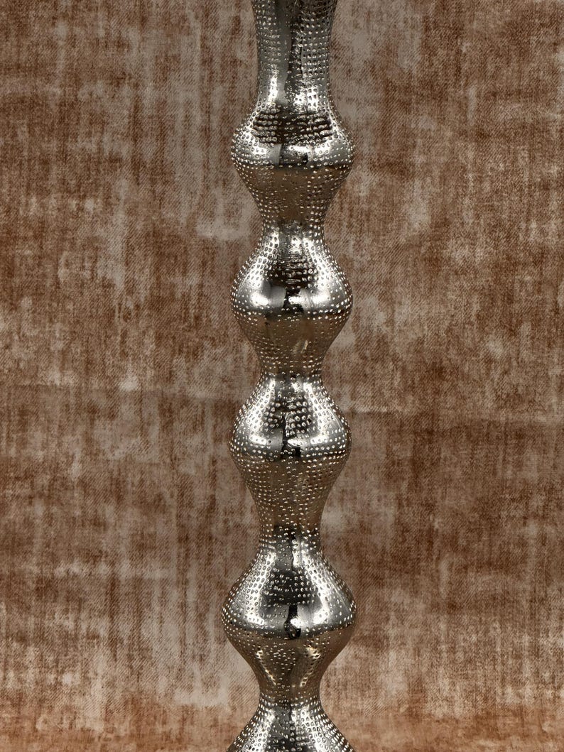 Pu&ograve; includere: Un oggetto decorativo alto in metallo argentato con una superficie strutturata e puntinata. L'oggetto presenta una serie di sezioni bulbose e arrotondate impilate l'una sull'altra, creando una forma allungata unica. La superficie riflettente cattura la luce.