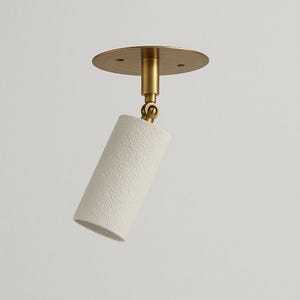 Op de afbeelding: Een moderne plafondspot met een getextureerde witte cilindrische kap en een messingkleurige montageplaat en steel. De spot is verstelbaar voor gerichte verlichting.