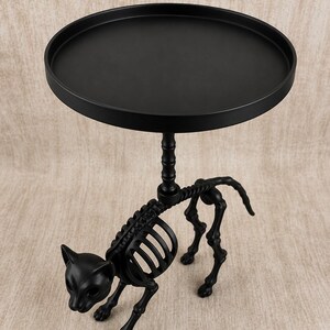 May include: Black metal side table with a round top and a cat skeleton base. The table has a matte finish and is designed with a Halloween or gothic theme. The cat skeleton is detailed and adds a unique decorative element.