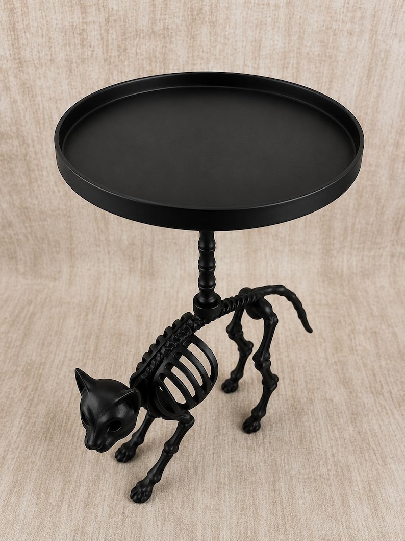 May include: Black metal side table with a round top and a cat skeleton base. The table has a matte finish and is designed with a Halloween or gothic theme. The cat skeleton is detailed and adds a unique decorative element.