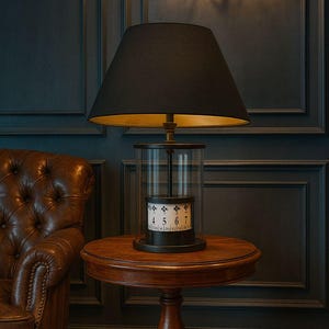 May include: A black table lamp with a gold interior shade sits on a small wooden table. The lamp has a clear glass cylinder base with a rotating numbered dial. A brown leather armchair is to the left, and a gold sconce is on the dark blue wall.