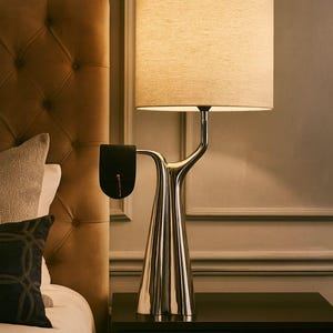 May include: A modern bedside lamp with a silver base and a beige fabric shade. The lamp has a unique, curved design and sits on a black nightstand. A black accessory with a red accent hangs from the lamp. The background includes a tufted headboard.