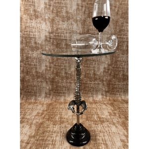 May include: A unique side table with a clear glass top and a silver-colored spine-shaped support. The base is black and round. Two wine glasses, one upright and one on its side, are on the table, filled with red wine.