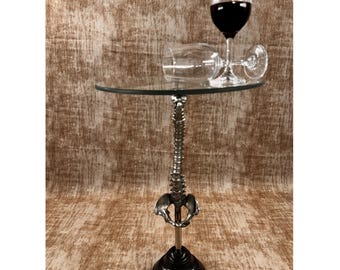May include: A distinctive side table featuring a clear glass top and a silver-coloured spine-shaped support. The base is black and round. Two wine glasses, one upright and one on its side, are on the table, filled with red wine.