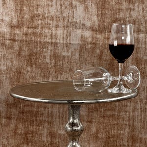 Pu&ograve; includere: Un tavolino d'argento con piano strutturato e base decorativa. Due calici, uno in posizione verticale e pieno di vino rosso, l'altro appoggiato su un lato. Lo sfondo &egrave; una parete marrone strutturata.