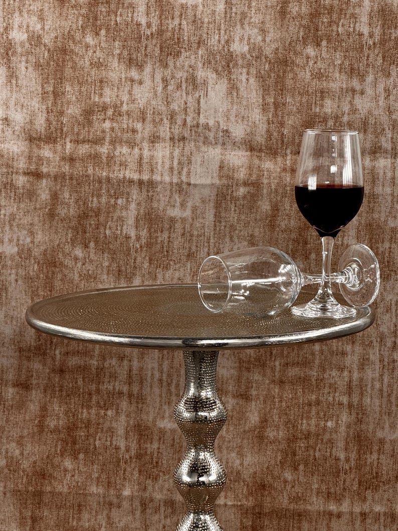 Pu&ograve; includere: Un tavolino d'argento con piano strutturato e base decorativa. Due calici, uno in posizione verticale e pieno di vino rosso, l'altro appoggiato su un lato. Lo sfondo &egrave; una parete marrone strutturata.