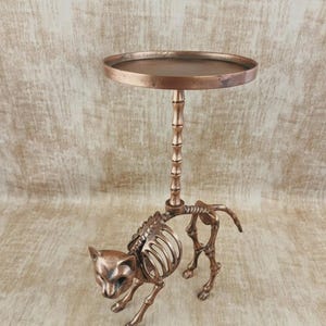 May include: A bronze-colored cat skeleton table with a round top. The cat skeleton is in a walking pose, with the table's stem rising from its back. The table is a unique decorative item.