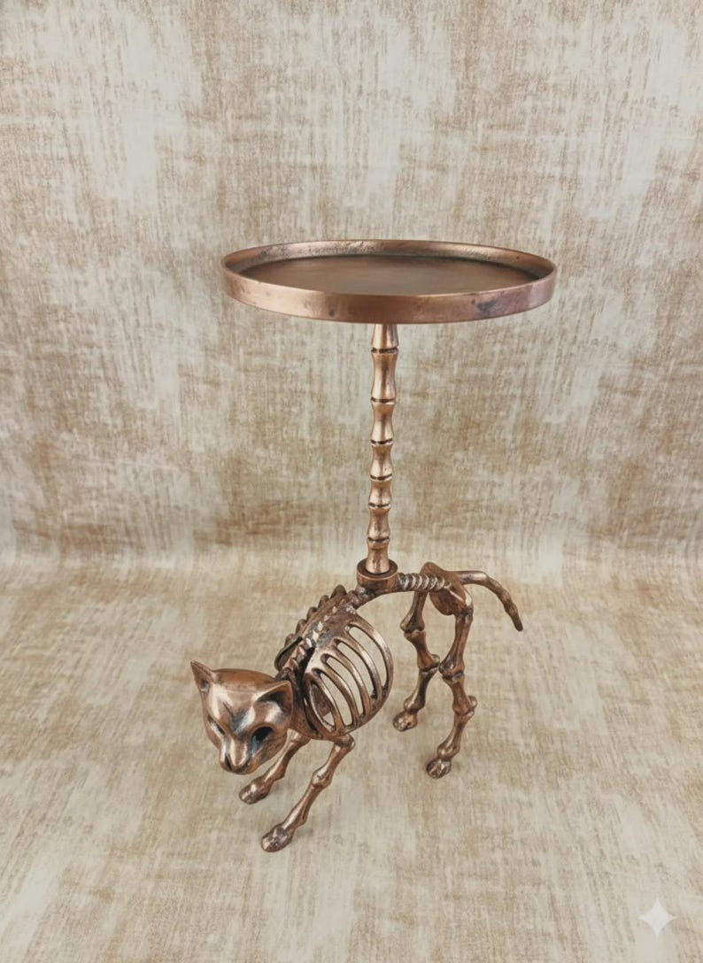 May include: A bronze-colored cat skeleton table with a round top. The cat skeleton is in a walking pose, with the table's stem rising from its back. The table is a unique decorative item.