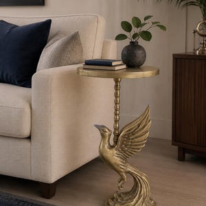 Puede incluir: Una mesa auxiliar de metal en tono dorado con un tablero circular sostenido por una columna central y una base esculpida con la forma de un fénix con las alas extendidas. La mesa tiene un acabado metálico desgastado y una base redonda.