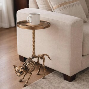 May include: A unique side table with a cat skeleton base and a round, antique brass tray top. A white mug with a gold rim sits on the tray. The table is next to a beige sofa, creating a stylish accent piece for a living room.