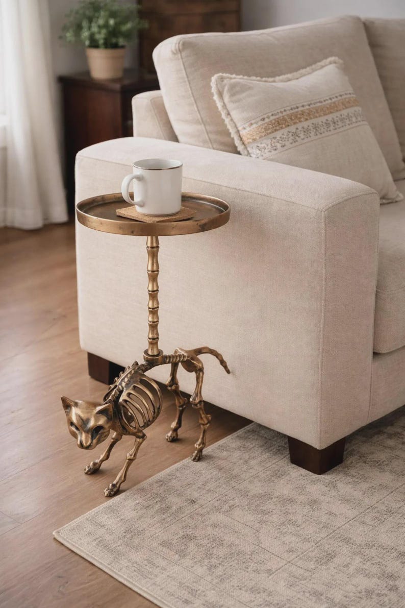 May include: A unique side table with a cat skeleton base and a round, antique brass tray top. A white mug with a gold rim sits on the tray. The table is next to a beige sofa, creating a stylish accent piece for a living room.