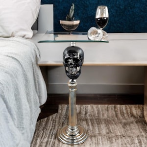 May include: A decorative side table with a black skull design. The table features a silver-colored pedestal base and a clear glass top. The table is placed next to a bed with white bedding.