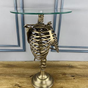 May include: A unique side table featuring a bronze-colored skeleton torso base supporting a round glass top. The table has a detailed rib cage and spine design, adding a gothic or anatomical aesthetic. The table stands on a round base.