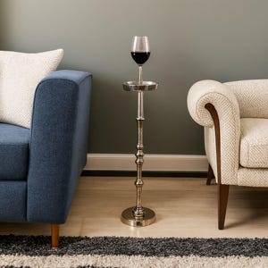 May include: A silver-colored wine glass holder with a glass of red wine. The holder has a round base, a slender, decorative stem, and a small tray to hold the wine glass. The holder is placed between a blue sofa and a cream-colored armchair.