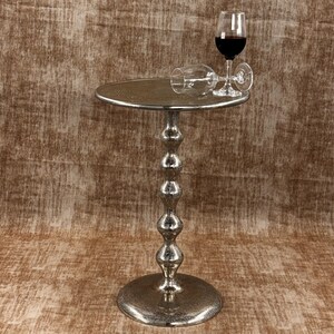 Pu&ograve; includere: Un tavolino argentato con perline e piano rotondo. Due calici, uno in posizione verticale con vino rosso e uno sdraiato, poggiano sul tavolo. Il tavolo ha una superficie strutturata e un gambo decorativo con perline.