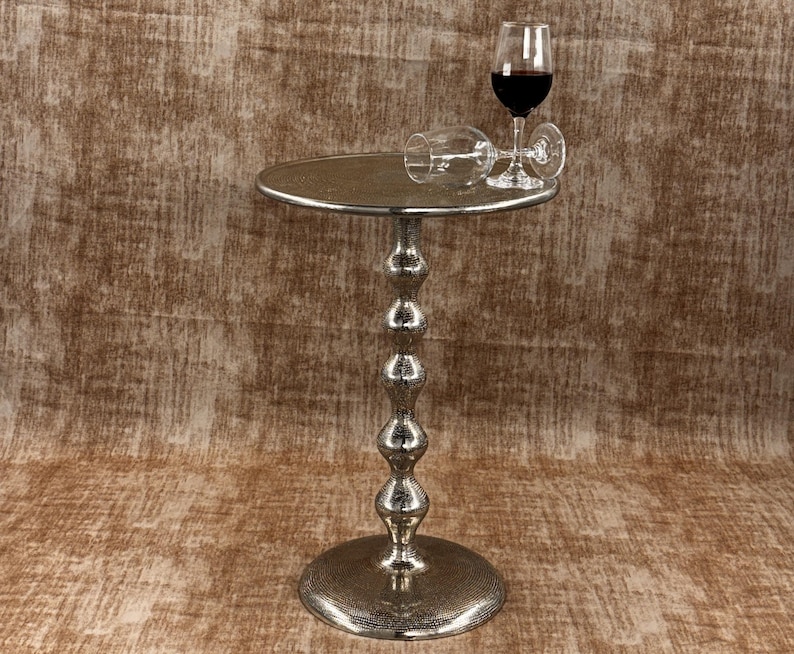 Pu&ograve; includere: Un tavolino argentato con perline e piano rotondo. Due calici, uno in posizione verticale con vino rosso e uno sdraiato, poggiano sul tavolo. Il tavolo ha una superficie strutturata e un gambo decorativo con perline.