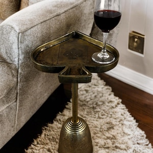 Puede incluir: Mesa auxiliar decorativa de latón antiguo con forma de pica. La mesa tiene un tallo largo y una base en forma de campana. Una copa de vino tinto descansa sobre la mesa. La mesa está colocada sobre una alfombra peluda.