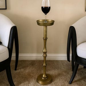 May include: A tall, antique gold-colored wine stand with a glass of red wine. The stand has a round base, a slender stem with decorative elements, and a small tray to hold the wine glass. The stand is approximately 36 inches tall.