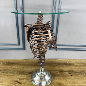 May include: A decorative side table with a skeleton torso base in copper tones, topped with a round glass surface. The base has a silver-coloured, pedestal-style foot. The table is a unique accent piece.