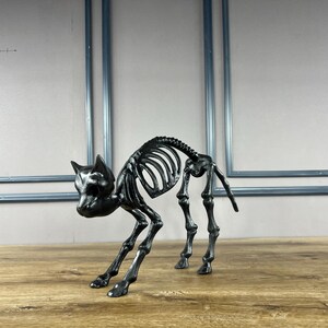 May include: A black cat skeleton ornament, ideal for Halloween. The cat is depicted in a crouching stance, with its rib cage and spine visible. The ornament is made of a dark, possibly metallic material.