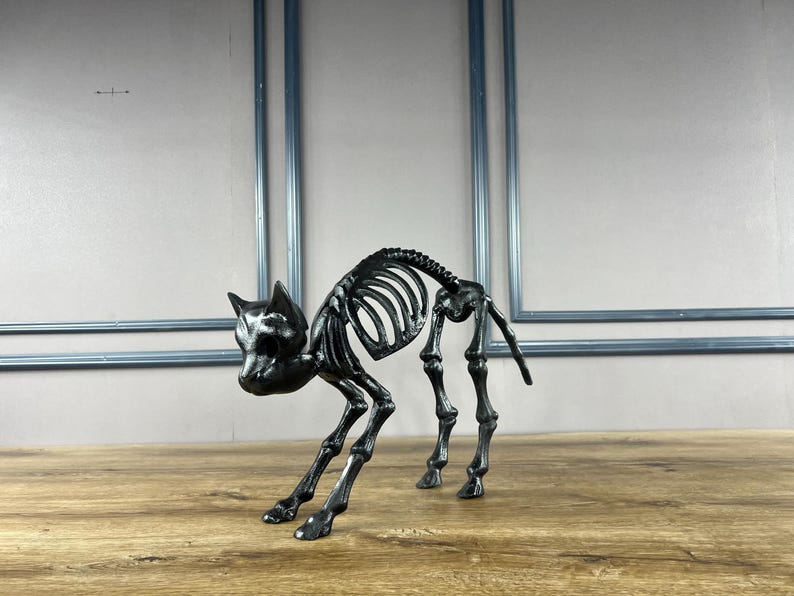 May include: A black cat skeleton ornament, ideal for Halloween. The cat is depicted in a crouching stance, with its rib cage and spine visible. The ornament is made of a dark, possibly metallic material.