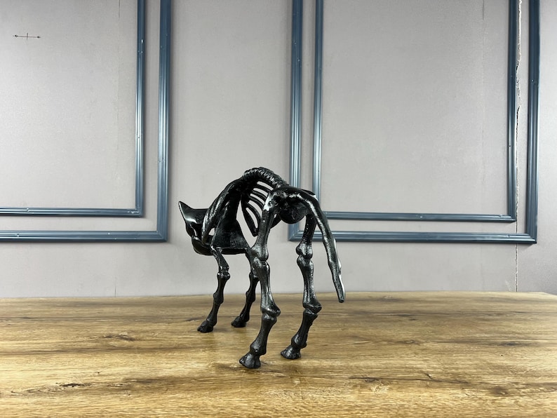 May include: A black cat skeleton sculpture with a detailed rib cage and leg bones. The sculpture is positioned on a wooden surface, with a light grey wall and blue framed rectangles in the background. The cat is in a crouching position.