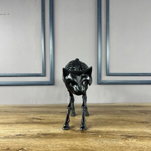 May include: A black cat skeleton sculpture with detailed rib cage and facial features. The cat is posed in a walking stance on a wooden surface. Two blue framed rectangles are in the background.