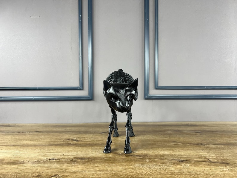 May include: A black cat skeleton sculpture with detailed rib cage and facial features. The cat is posed in a walking stance on a wooden surface. Two blue framed rectangles are in the background.
