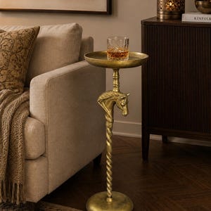 May include: A gold-toned metal side table featuring a circular tabletop, a central pedestal with a twisted design, and a sculpted horse head detail. The table stands on a round base and holds a clear glass tumbler containing an amber liquid.