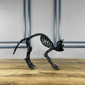 May include: A black cat skeleton ornament, depicted in a stalking pose. The sculpture is crafted from a dark material and is set against a plain backdrop. The cat's rib cage and spine are clearly visible.