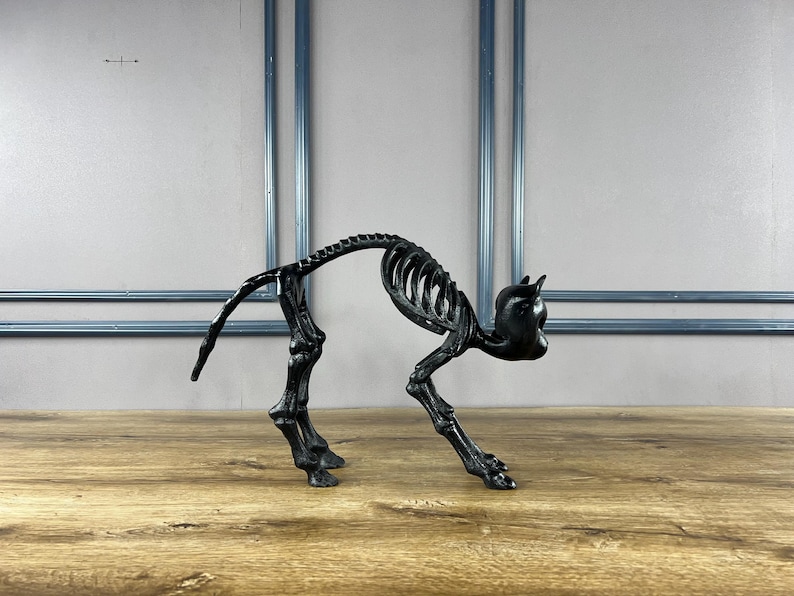 May include: A black cat skeleton ornament, depicted in a stalking pose. The sculpture is crafted from a dark material and is set against a plain backdrop. The cat's rib cage and spine are clearly visible.