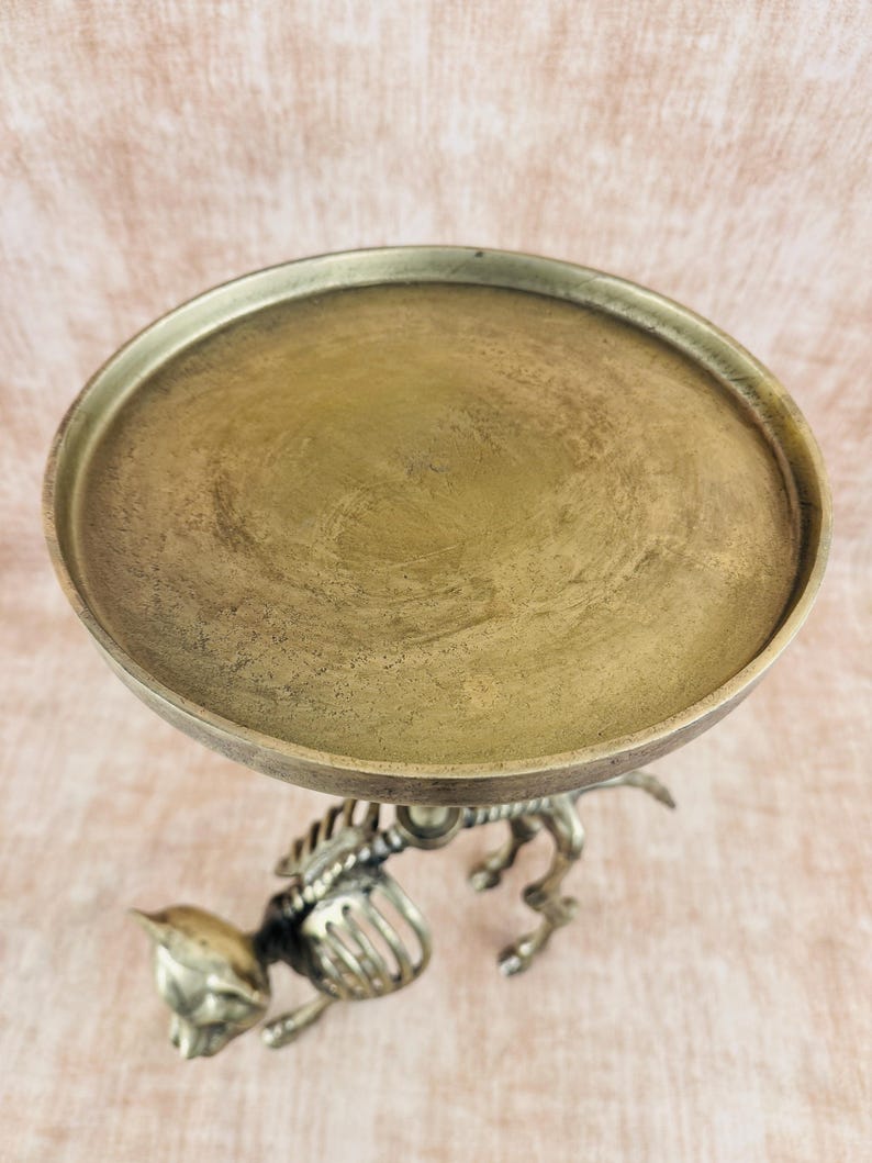 May include: A decorative, antique-style metal stand with a round, brass-colored tray. The stand is supported by a detailed, silver-colored cat skeleton design. The tray is approximately 8 inches in diameter.