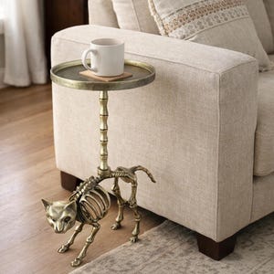May include: A unique side table with a cat skeleton base and a round, antique brass-colored tabletop. A white mug rests on a coaster on the table. The table is next to a beige sofa.