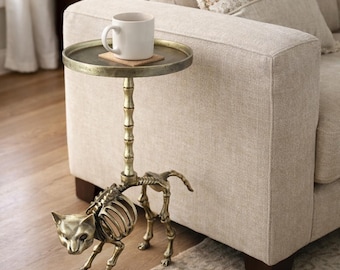 May include: A decorative side table featuring a cat skeleton base and a circular, antique brass-coloured tabletop. A white mug sits on a coaster. The table is beside a beige sofa.