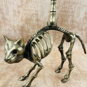 May include: A brass cat skeleton figurine with a candle holder. The cat is posed in a walking position, with detailed rib cage and leg bones. The candle holder is a vertical rod extending from the cat's back. The figurine has a vintage, antique appearance.