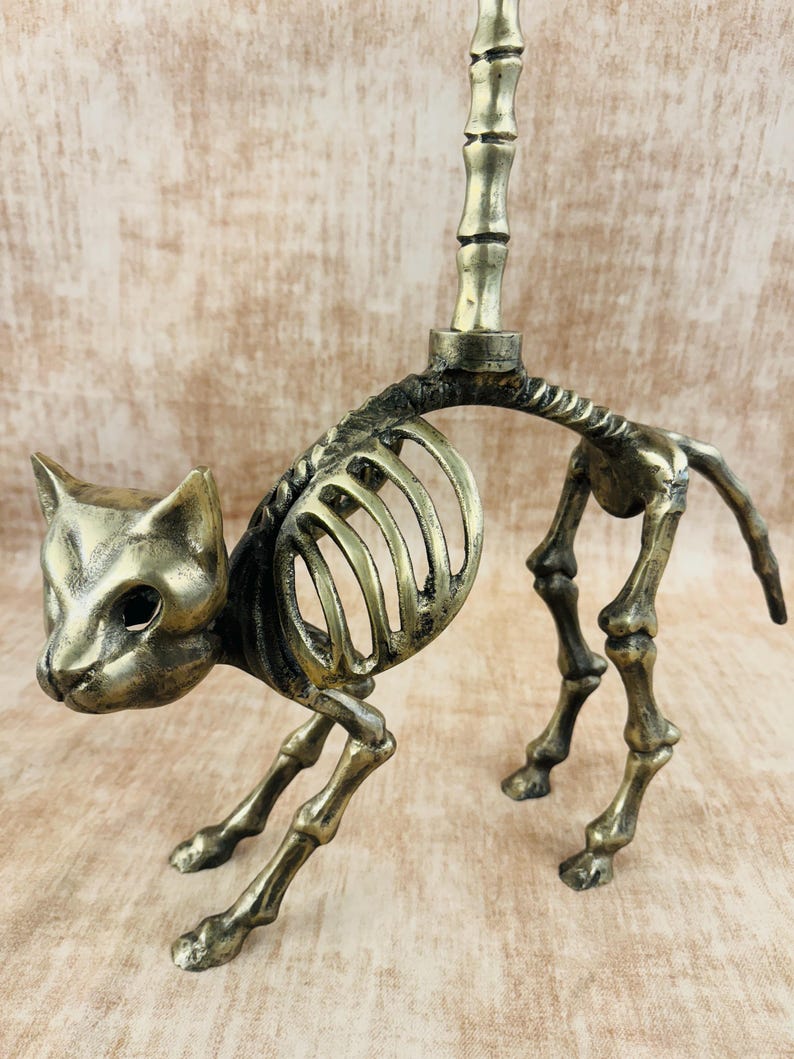May include: A brass cat skeleton figurine with a candle holder. The cat is posed in a walking position, with detailed rib cage and leg bones. The candle holder is a vertical rod extending from the cat's back. The figurine has a vintage, antique appearance.