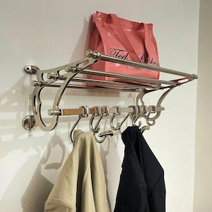 Può includere: Un attaccapanni a parete cromato con un ripiano superiore e ganci. Una borsa tote rosa è appoggiata sul ripiano. Due cappotti, uno beige e uno nero, sono appesi ai ganci.