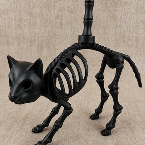 May include: A black cat skeleton figurine with a detailed rib cage, legs, and head. The cat is posed in a walking stance, with a vertical pole extending from its back. The figurine is made of a matte black material.