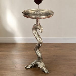 Puede incluir: Una mesa auxiliar de tono plateado con una base esculpida en forma de serpiente enroscada, con un cuerpo texturizado de escamas y una base de tres patas. Una bandeja redonda y plana descansa sobre la serpiente, sosteniendo una copa de vino tinto.