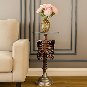 May include: A unique side table with a skeleton-themed design. The table features a bronze-colored rib cage and spine base, topped with a glass surface. A gold-colored heart-shaped vase holds pink flowers, adding a touch of elegance.