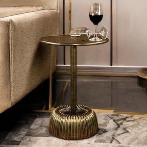 May include: A small, round side table with a gold finish. The table has a fluted base and a single central support. A wine glass with red wine and an empty wine glass are on the table. The table is next to a beige sofa.