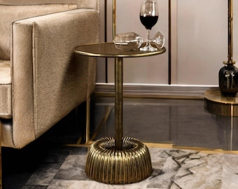 May include: A small, round side table with a gold finish. The table has a fluted base and a single central support. A wine glass with red wine and an empty wine glass are on the table. The table is next to a beige sofa.