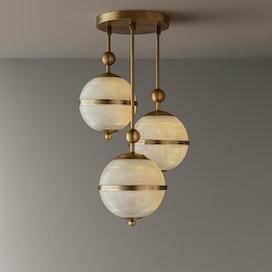Op de afbeelding: Een drielichts hanglamp met een messing afwerking. Het heeft drie bolvormige tinten met een getextureerde, gebroken witte uitstraling, geaccentueerd door messing banden. De lamp is opgehangen aan een ronde plafondbevestiging.