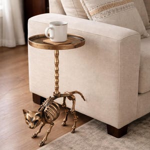 May include: A unique side table with a cat skeleton base and a round, brass-colored tabletop. A white mug sits on the table, which is next to a beige sofa. The table adds a touch of whimsy to the living room.