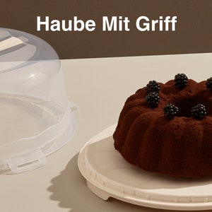 May include: A clear plastic cake dome with a handle, a white cake stand, and a chocolate cake with blackberries on top. The text "Haube Mit Griff" is written in white on a brown background.